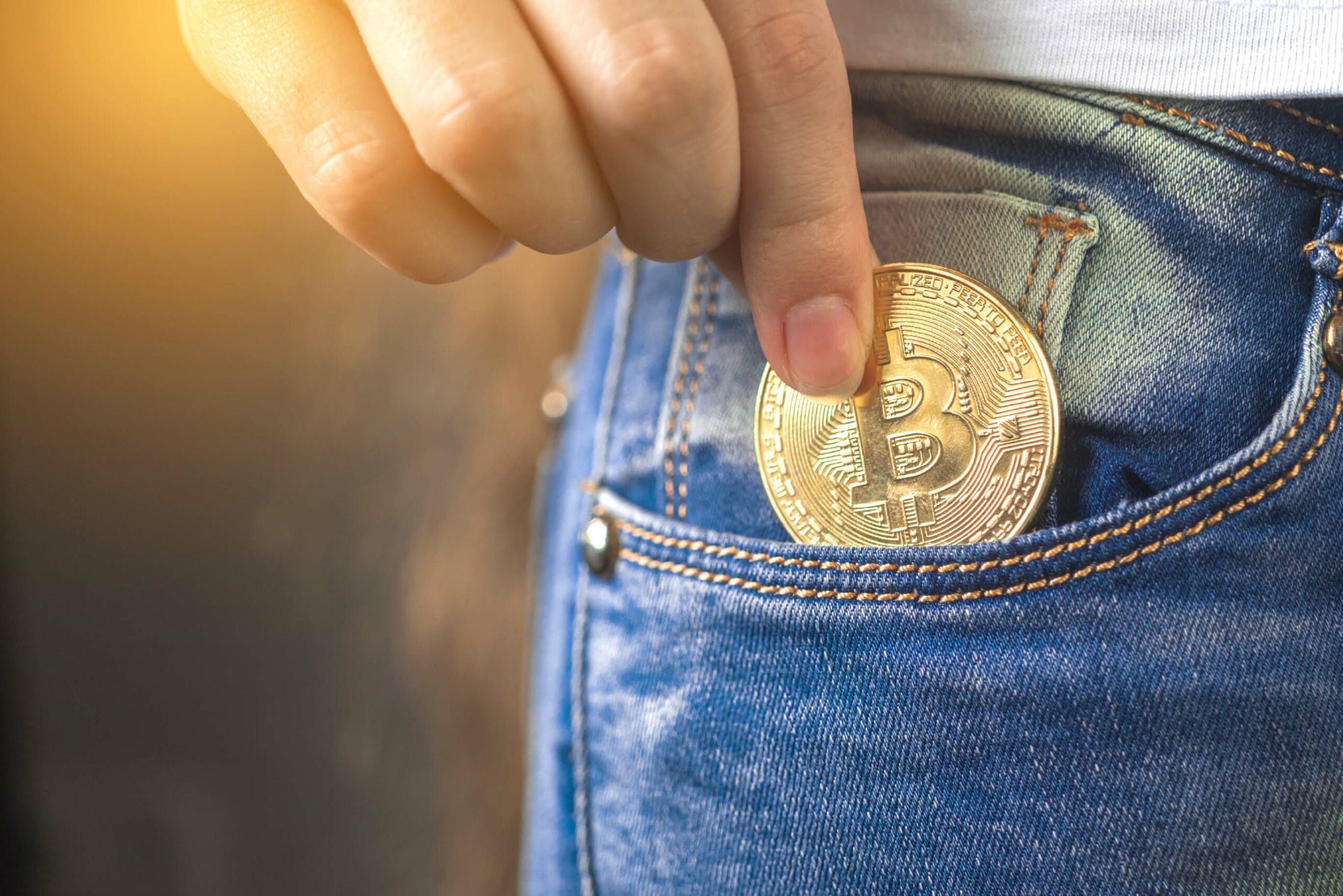 A close-up shot of someone placing a physical Bitcoin coin into the pocket of blue denim jeans. The coin is golden in color with the iconic Bitcoin 'B' symbol visible.