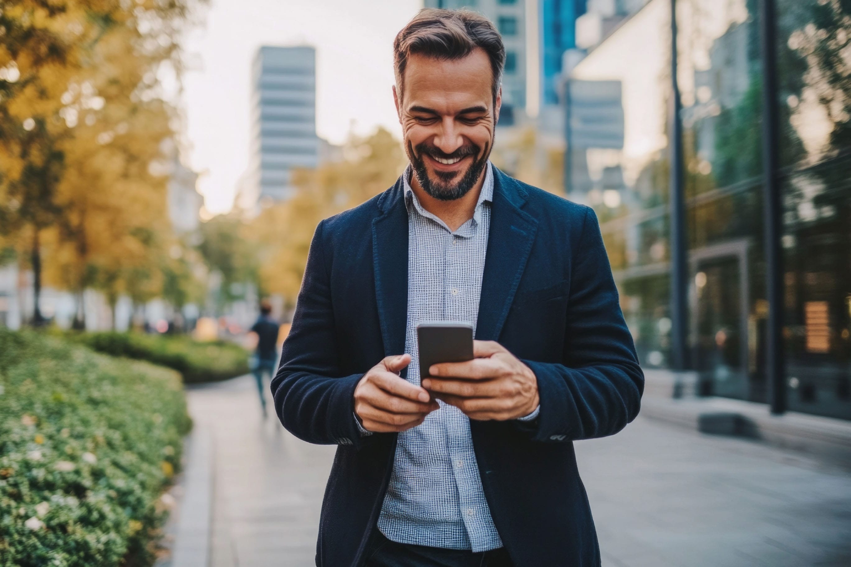 A smiling man with a beard, wearing a blazer over a patterned shirt, stands on an outdoor walkway looking down at his phone which he holds with both hands. The background shows trees, greenery, and modern city buildings.