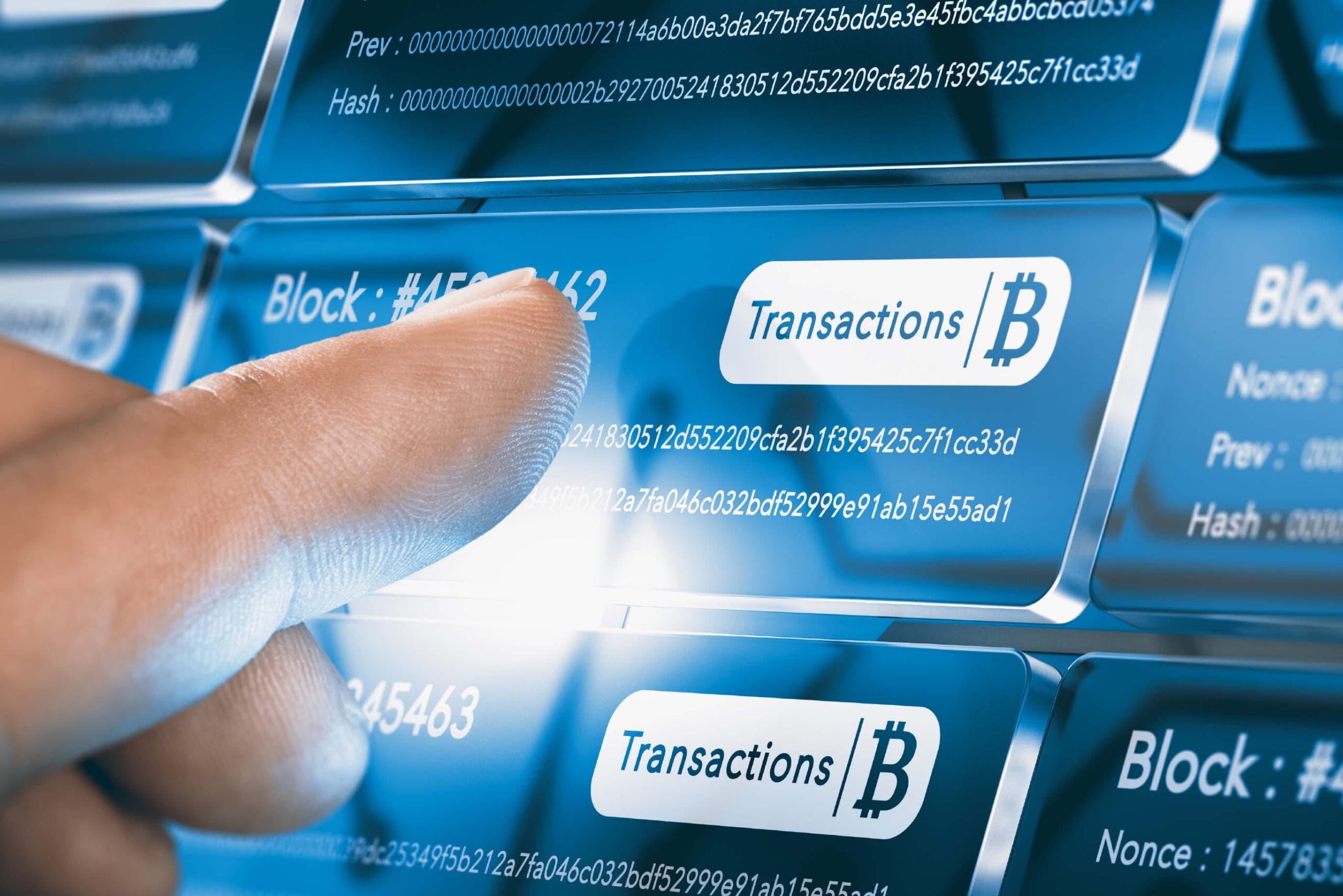 A finger pointing at a blockchain interface display showing Bitcoin transaction details in blue and white. The screen displays various technical information including block numbers, hash values, and transaction data with the Bitcoin symbol. Multiple rows of transaction information are visible with labels for "Block," "Hash," "Prev," and "Nonce" values.