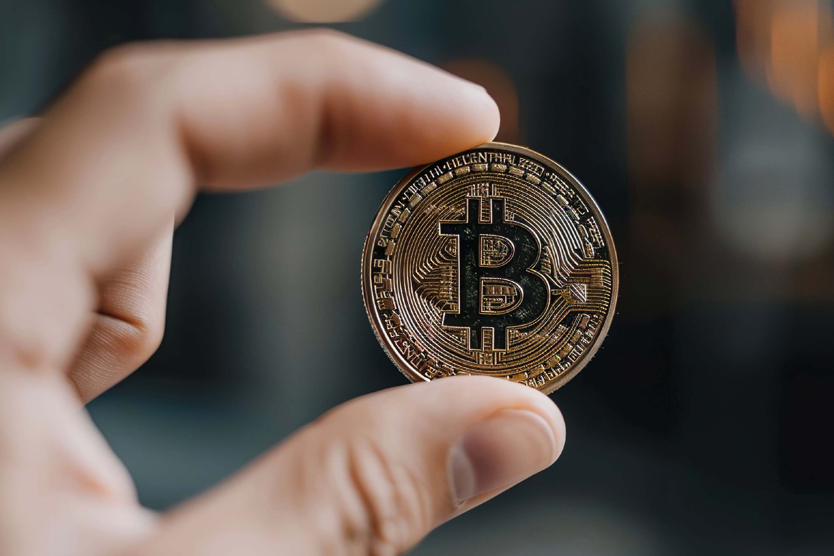Close-up photograph focusing on a person's thumb and index finger holding a detailed, physical Bitcoin coin. The coin is gold-colored with the Bitcoin 'B' symbol, and the background is softly blurred.