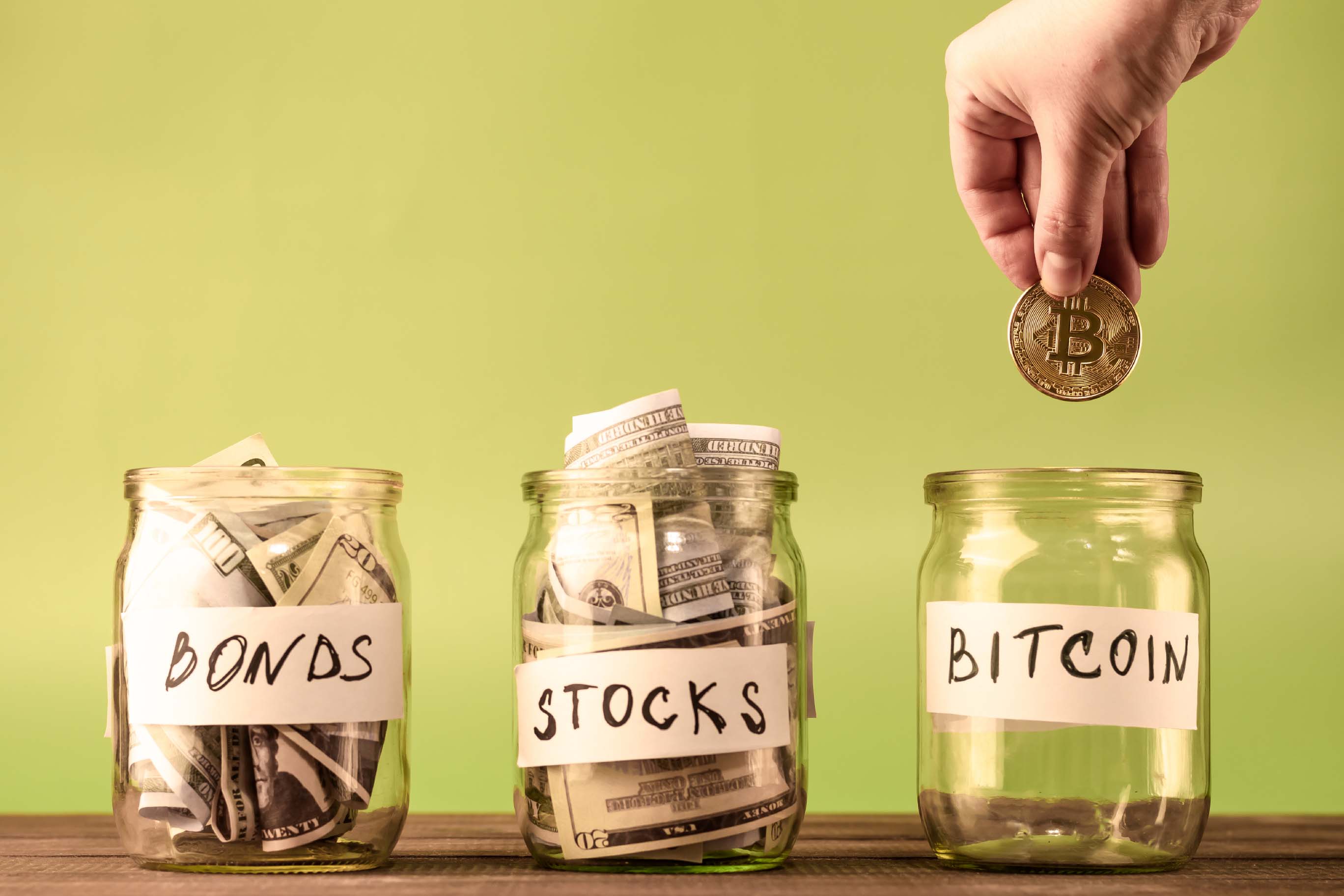 Three glass jars labeled 'BONDS', 'STOCKS', and 'BITCOIN'. The first two are filled with cash, the third is empty. A hand holds a Bitcoin coin above the 'BITCOIN' jar.
