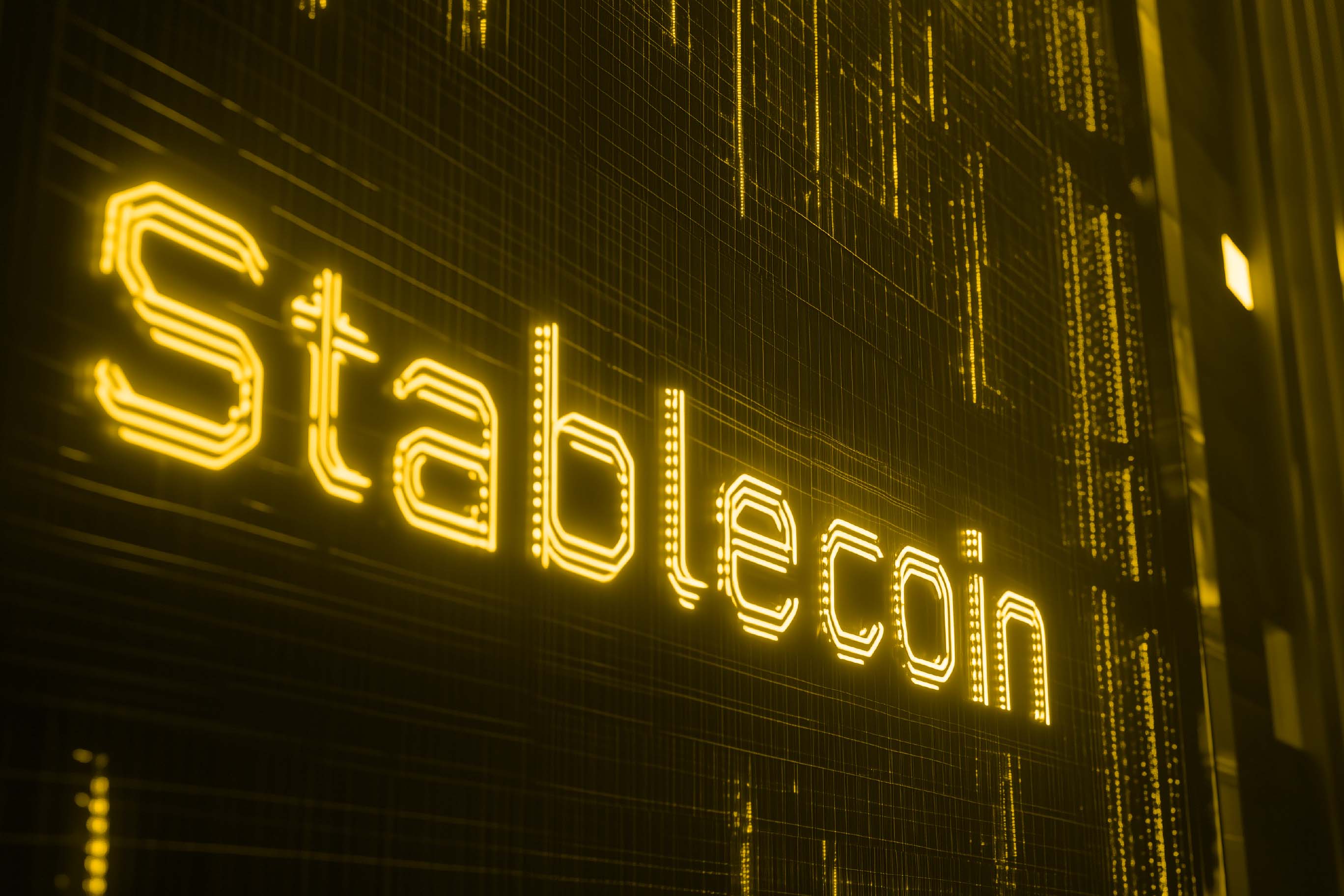 An angled view of the word 'Stablecoin' written in bright yellow, segmented letters that resemble a neon or digital display. The background is dark with faint grid lines and vertical data patterns.
