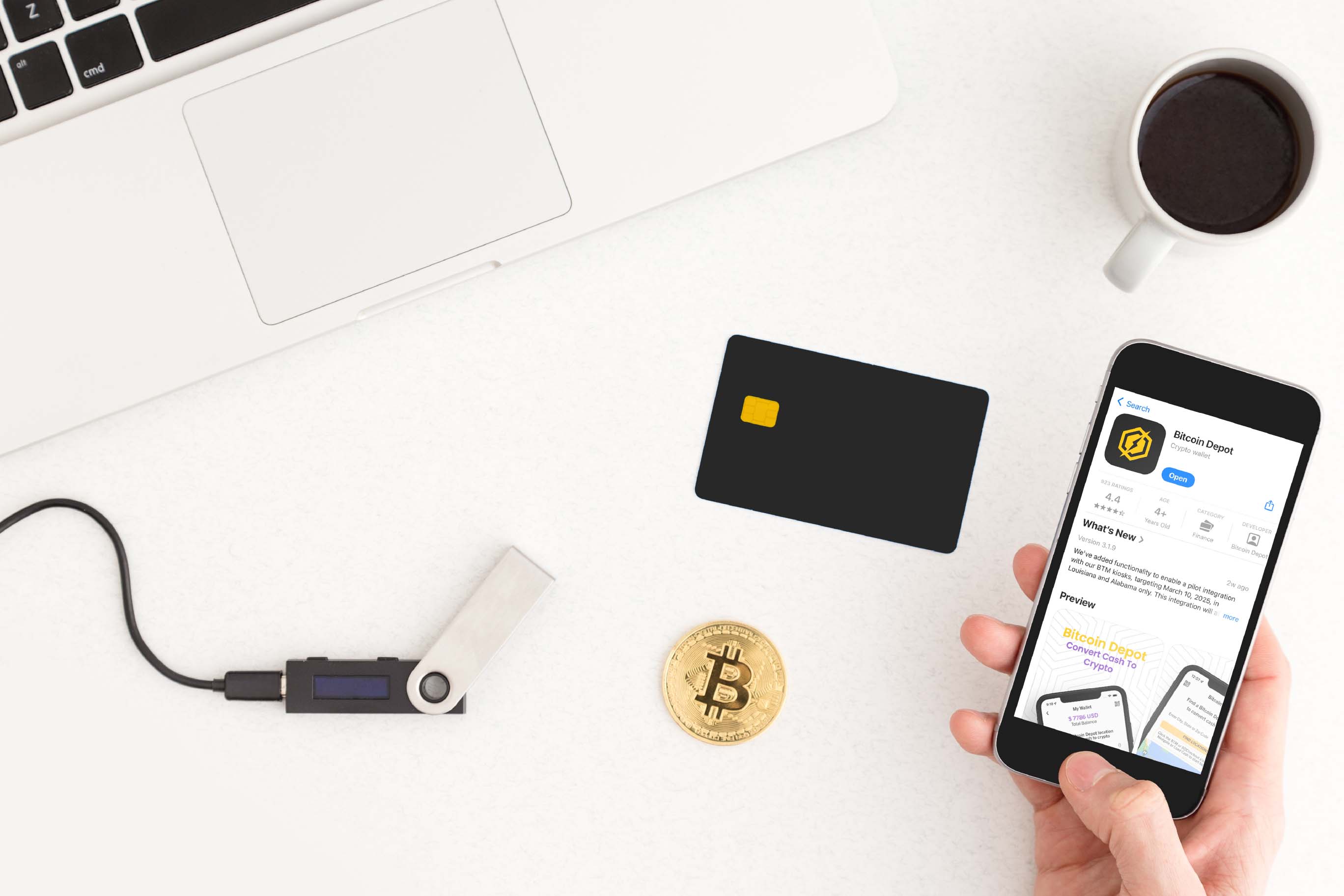 A flat lay on a white desk showing items related to cryptocurrency: a laptop, a hardware wallet, a physical Bitcoin coin, a debit card, and a hand holding a smartphone displaying the Bitcoin Depot app in the app store.