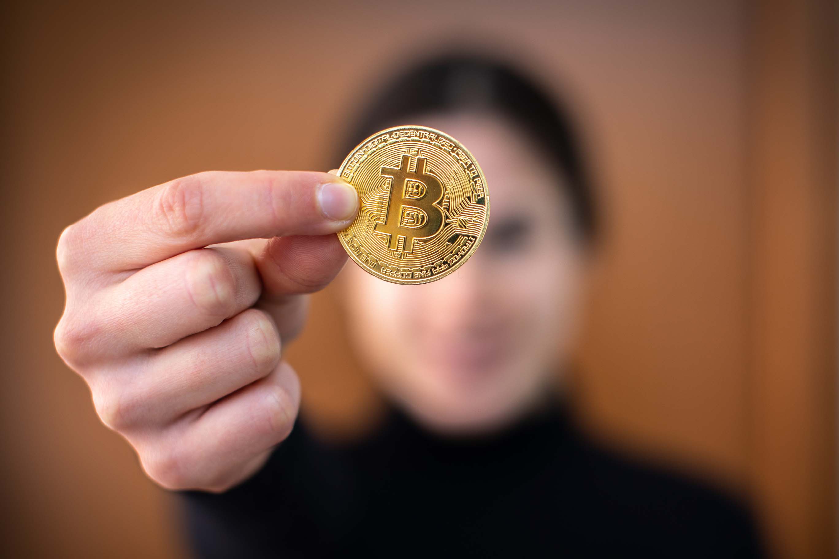 A hand, in sharp focus, extends forward to display a shiny, golden Bitcoin coin. The coin features the stylized "B" logo. In the blurred background, a person with dark hair is visible, looking towards the viewer.